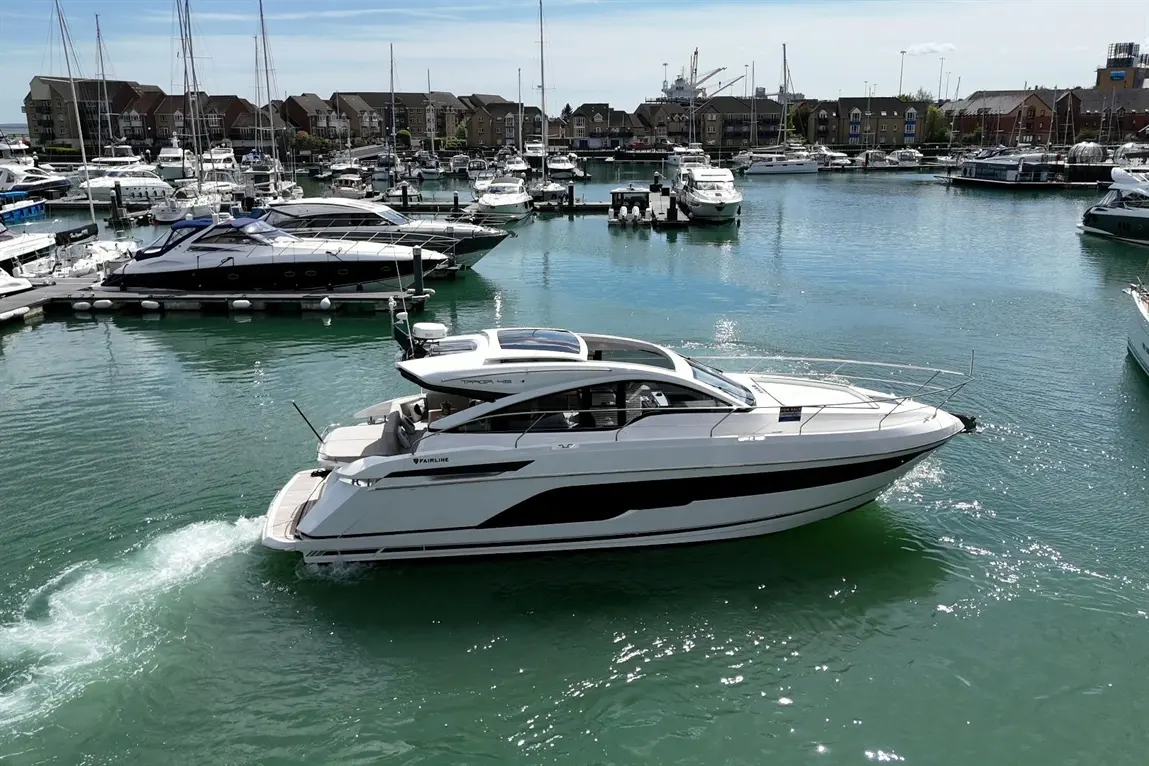 A modern Fairline motorboat navigates through a marina filled with yachts and waterfront buildings on a sunny day.