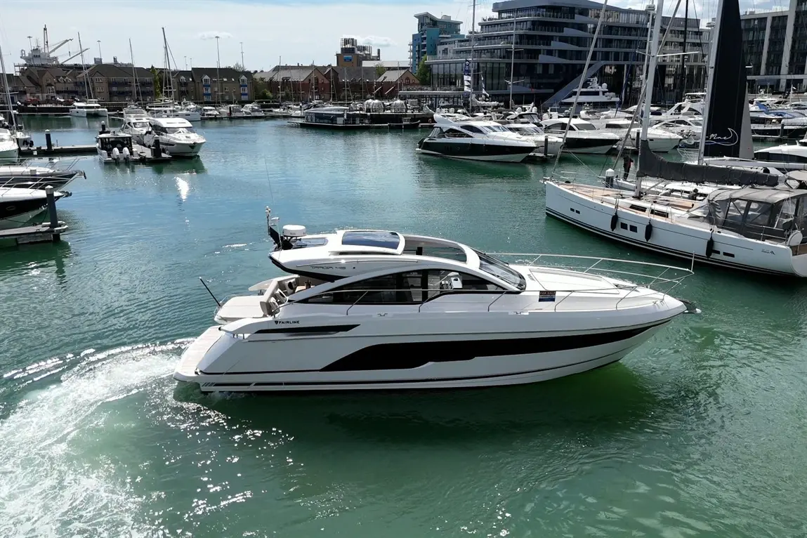 A sleek Fairline boat navigates the calm waters of a marina filled with various luxury yachts and boats.