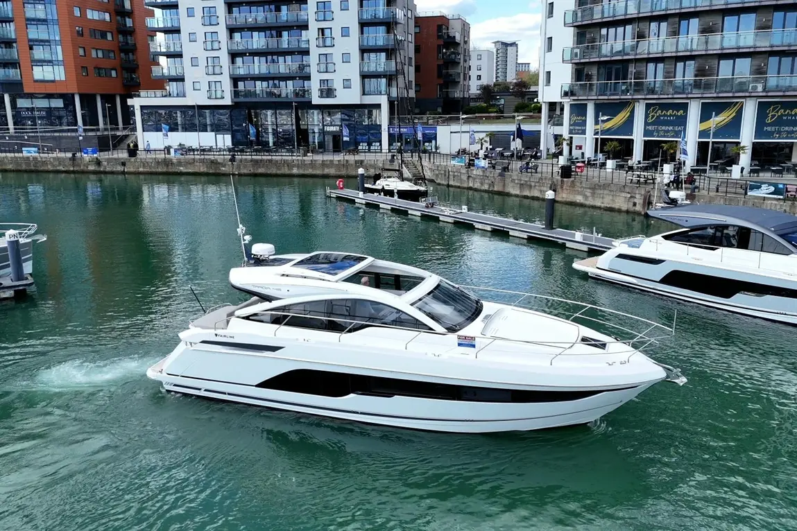 A sleek white motorboat, labelled 'Fairline', navigates greenish waters near a modern marina and waterfront buildings.
