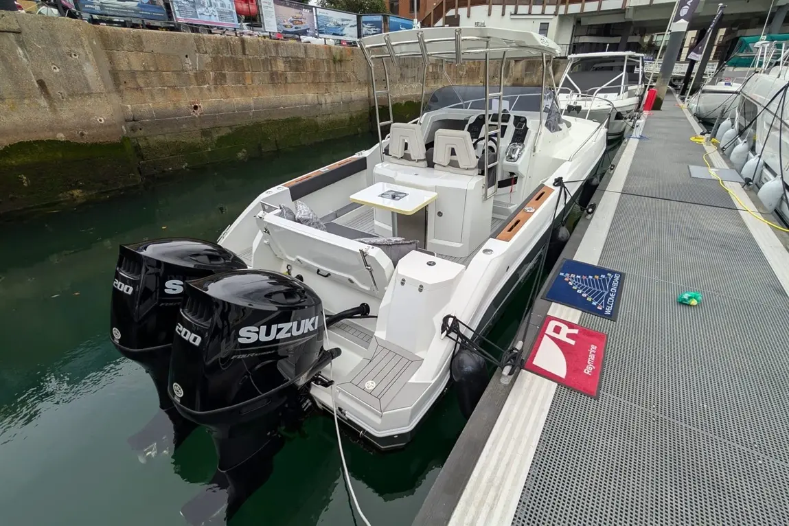 A white boat with twin Suzuki 200 engines is docked at a marina, featuring seating, a table, and nearby mats with logos.