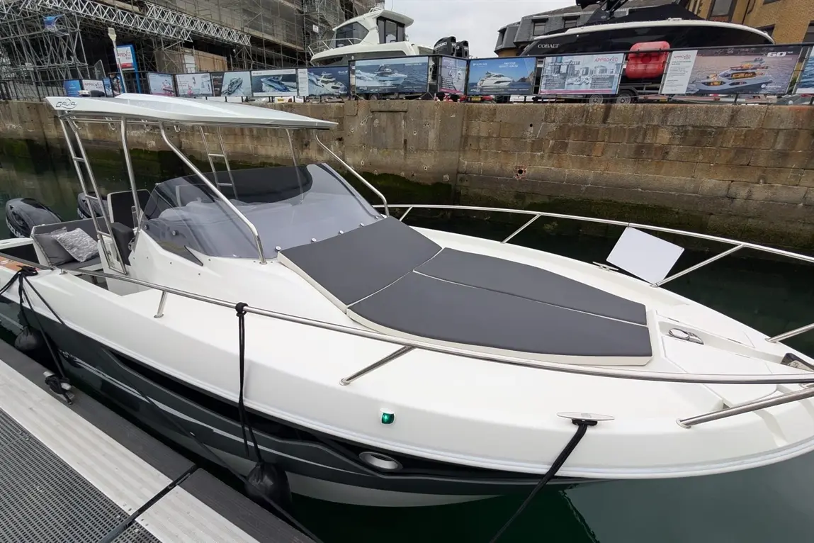 A sleek white boat with gray cushions docked at a marina, featuring a semi-open design and surrounding boats in the background.
