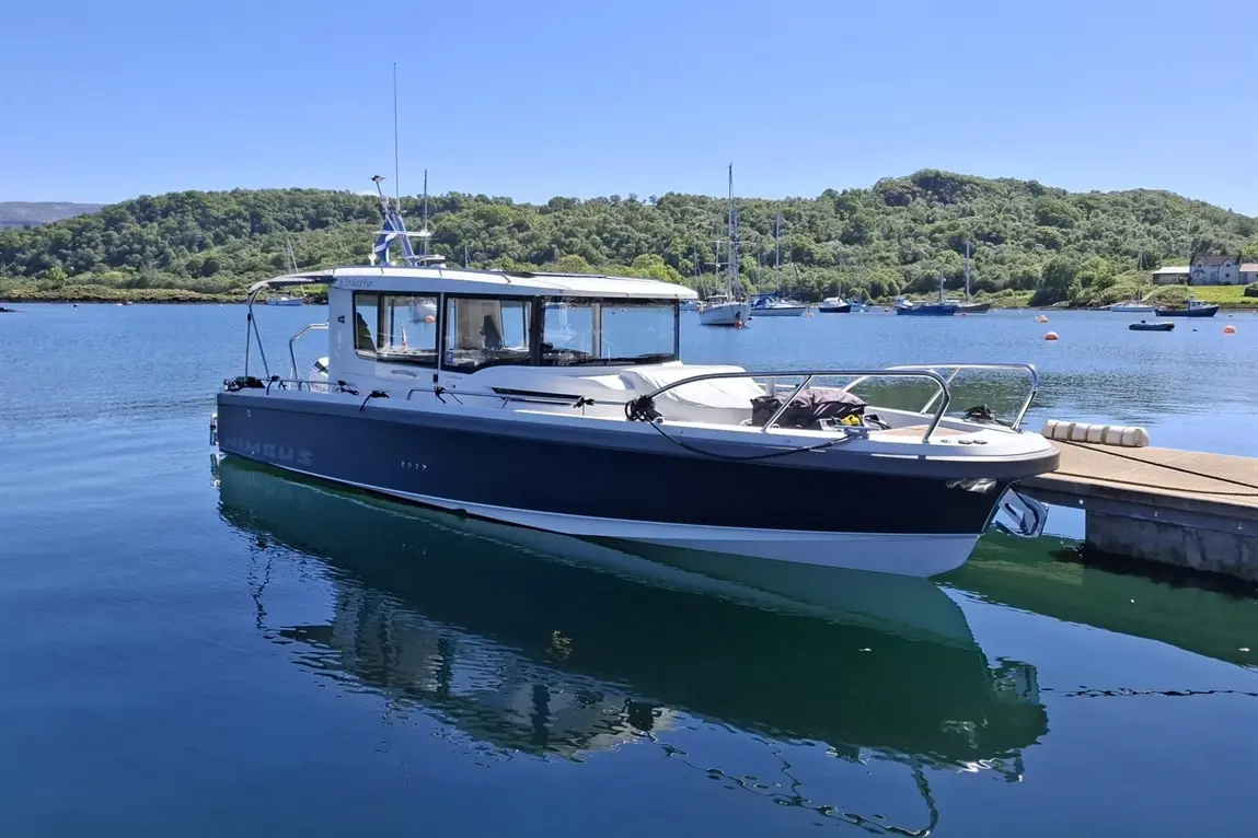 A Nimbus boat is anchored in calm water, surrounded by greenery and moored yachts under a clear blue sky.