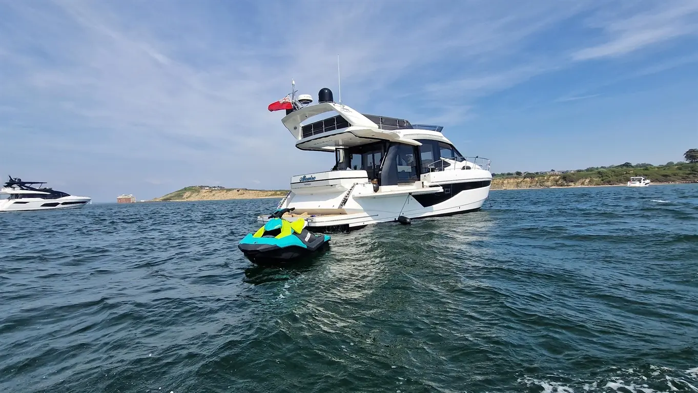 A luxury motor yacht sails in the sea, with a jet ski attached behind and a coastal landscape in the background.