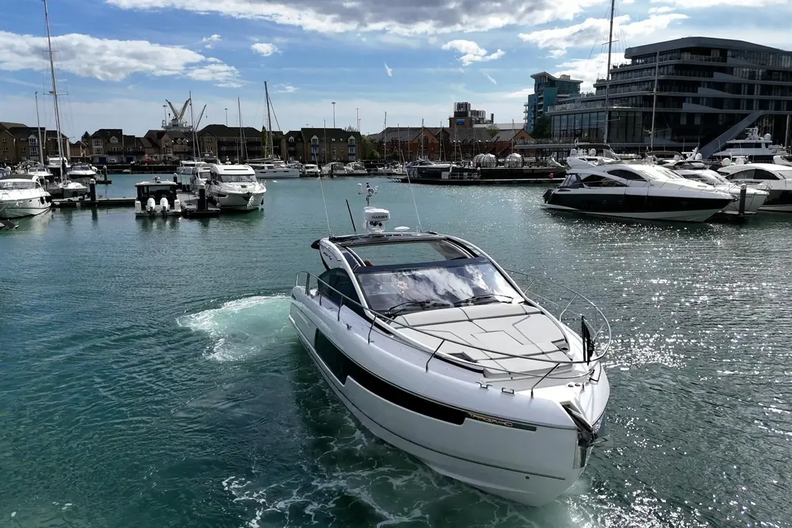 A sleek white motorboat navigates a marina filled with yachts under a bright blue sky with scattered clouds.