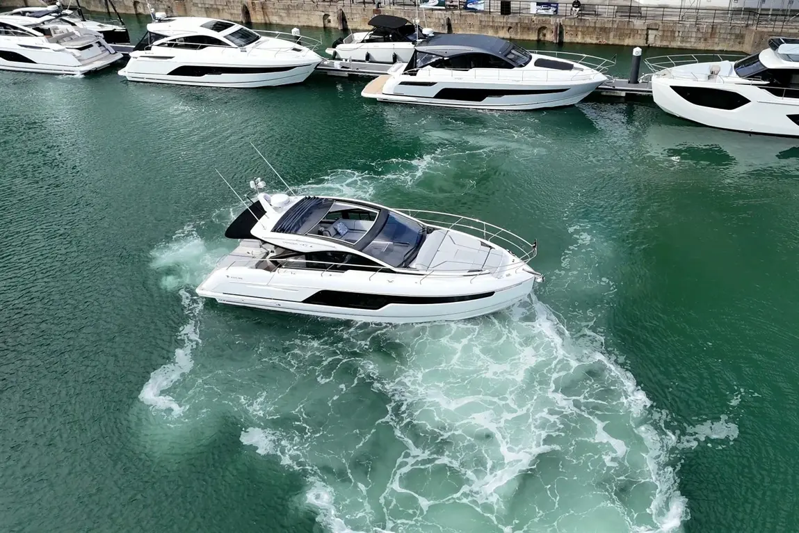A white motorboat navigates through clear green waters, surrounded by moored luxury boats in a marina setting.