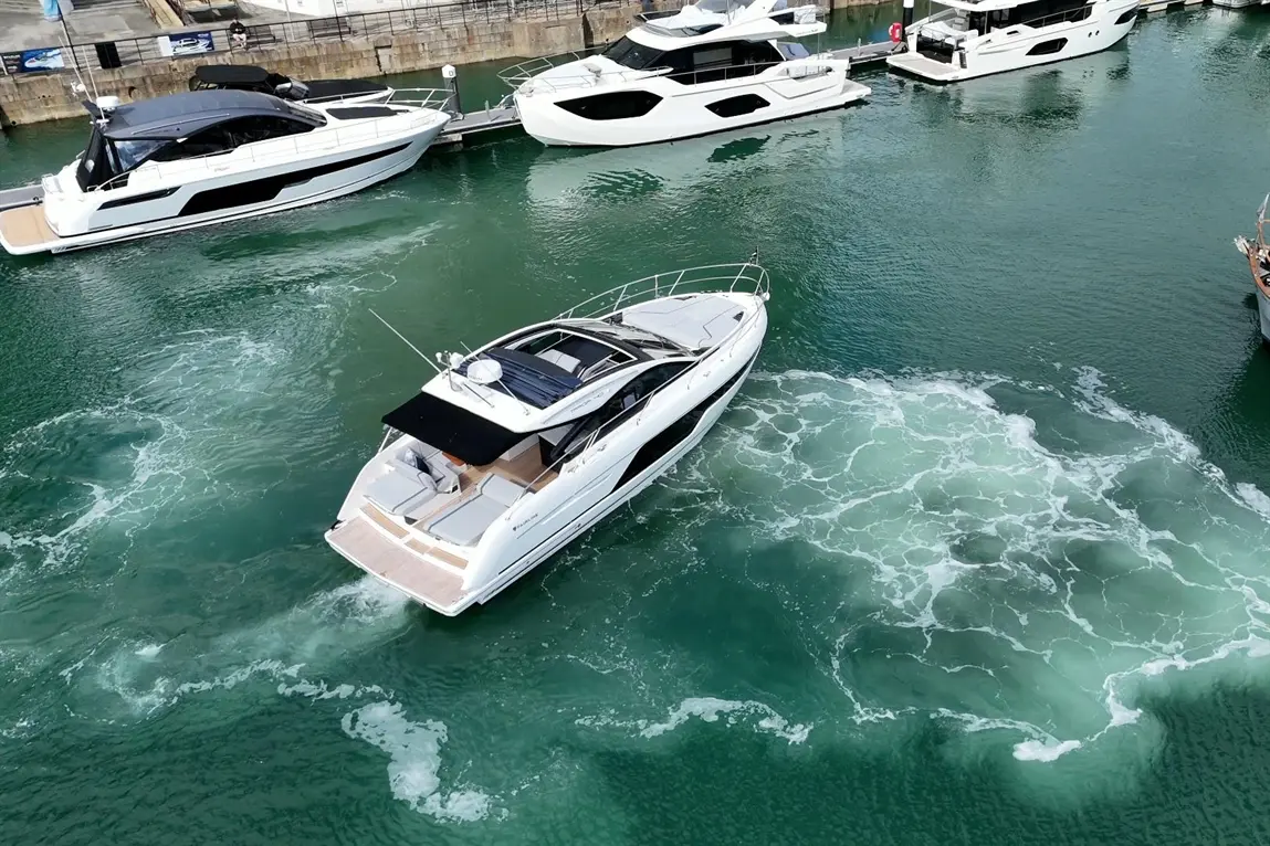 A sleek white motorboat navigates through emerald waters, leaving white foam trails near a marina with luxury yachts.