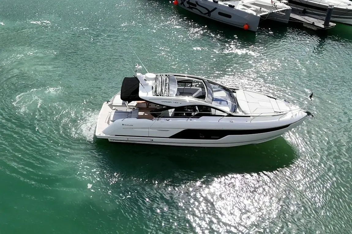 A sleek white motorboat glides through turquoise water, with sunlight glinting off the surface and nearby boats docked.