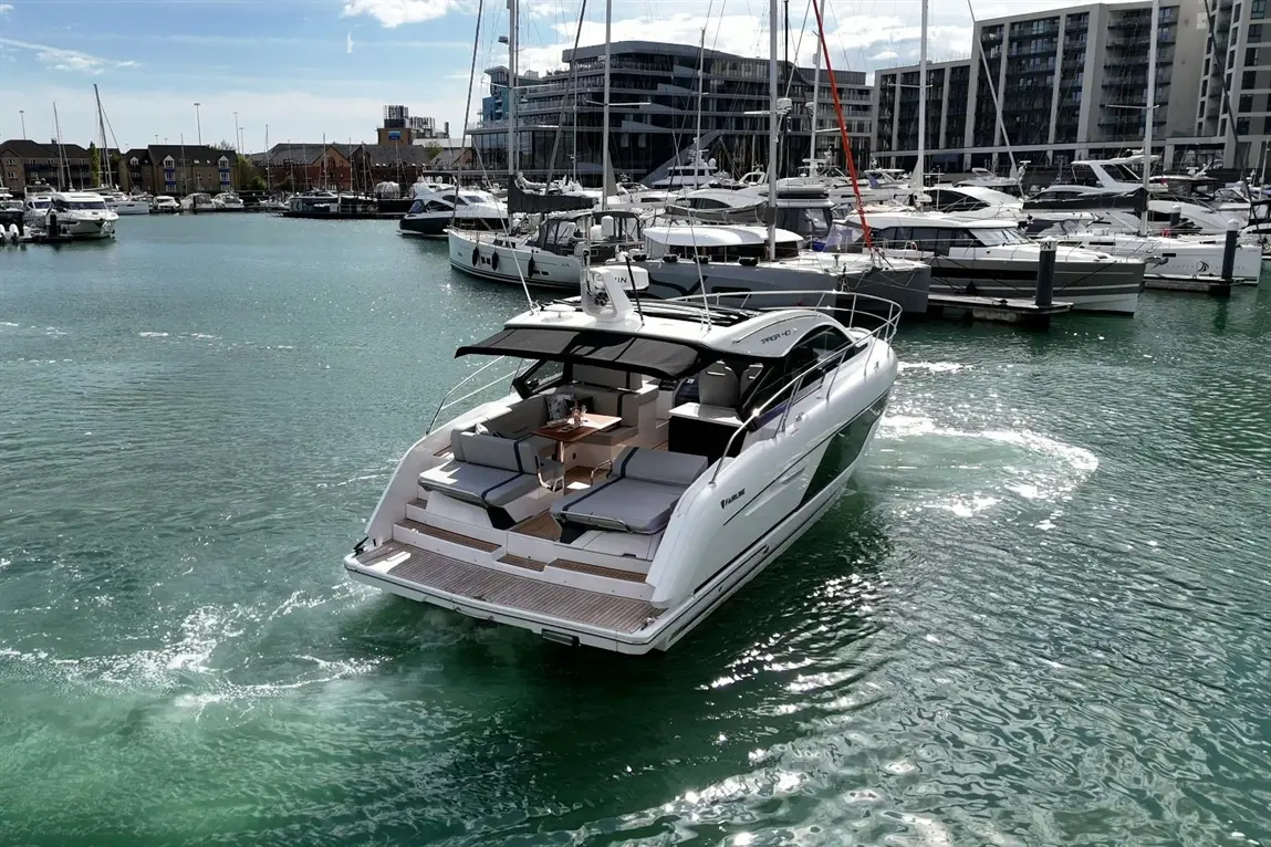 A sleek motorboat leaves the marina, surrounded by several boats, with modern buildings in the background and sparkling water.