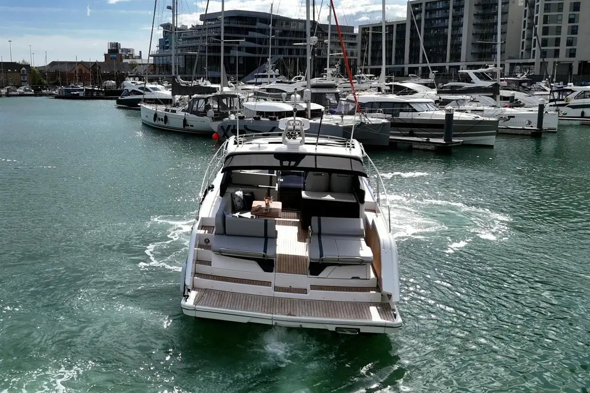 A luxury motorboat is leaving a marina, surrounded by various yachts and buildings on a sunny day.