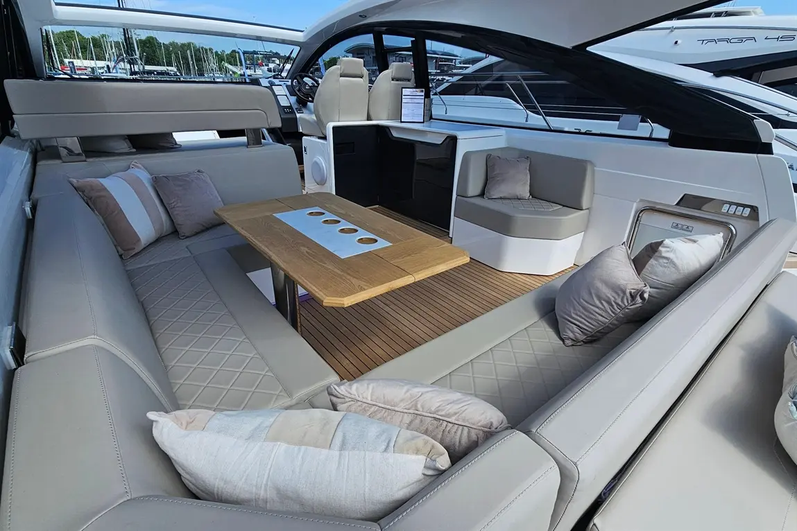 A modern boat interior featuring a comfy seating area, a wooden table with cup holders, and soft cushions, with yachts in the background.