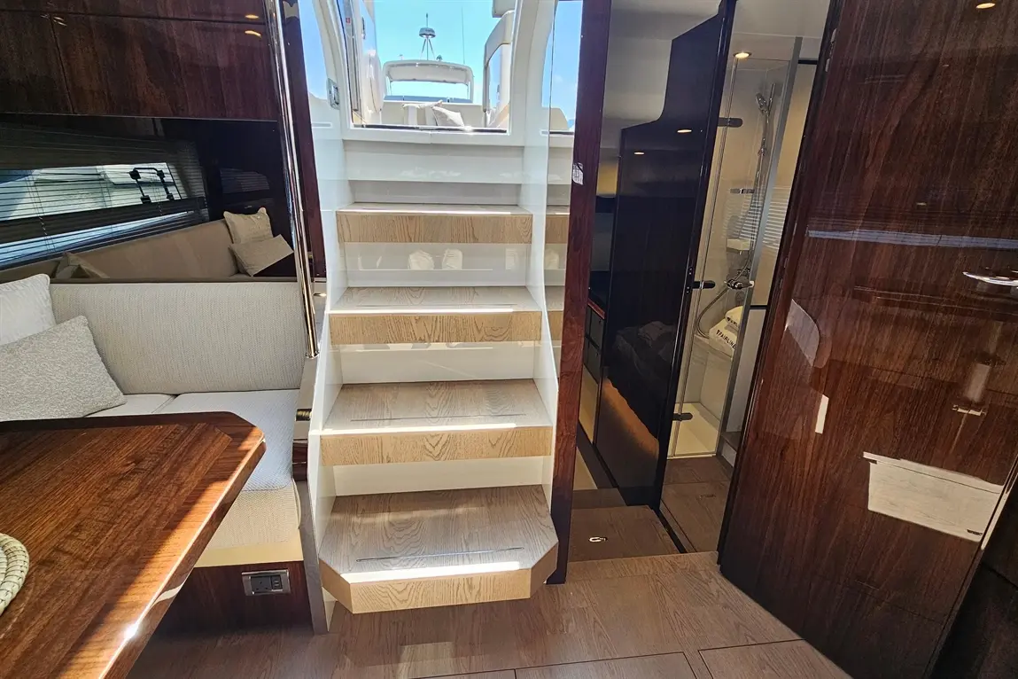 Wooden stairs illuminate the interior of a luxury yacht, with a spacious seating area and a doorway leading to the bathroom.