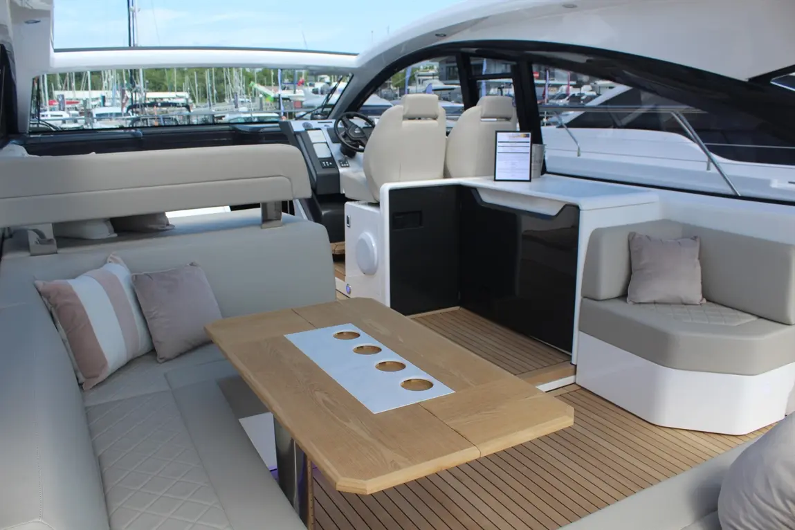Modern boat interior with beige seating, an oak table, and a control panel, overlooking a marina with moored yachts.