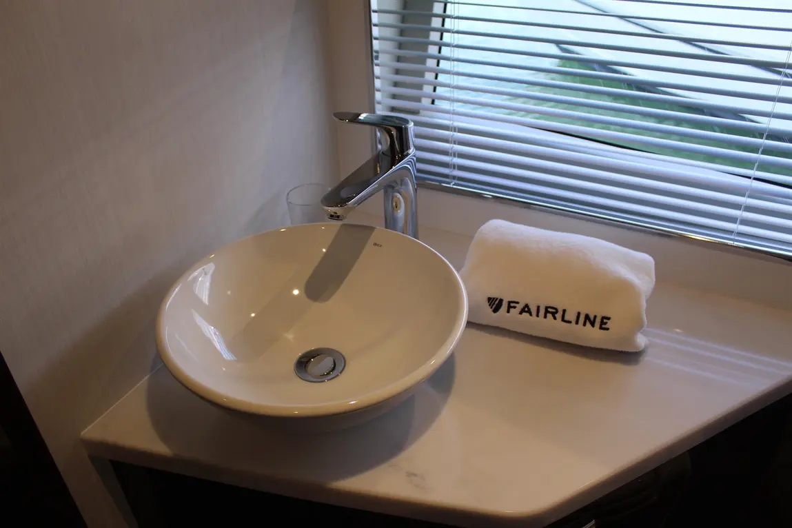 A modern white sink on a marble countertop with a chrome tap, next to a FAIRLINE towel and a glass. Blinds are partially open.
