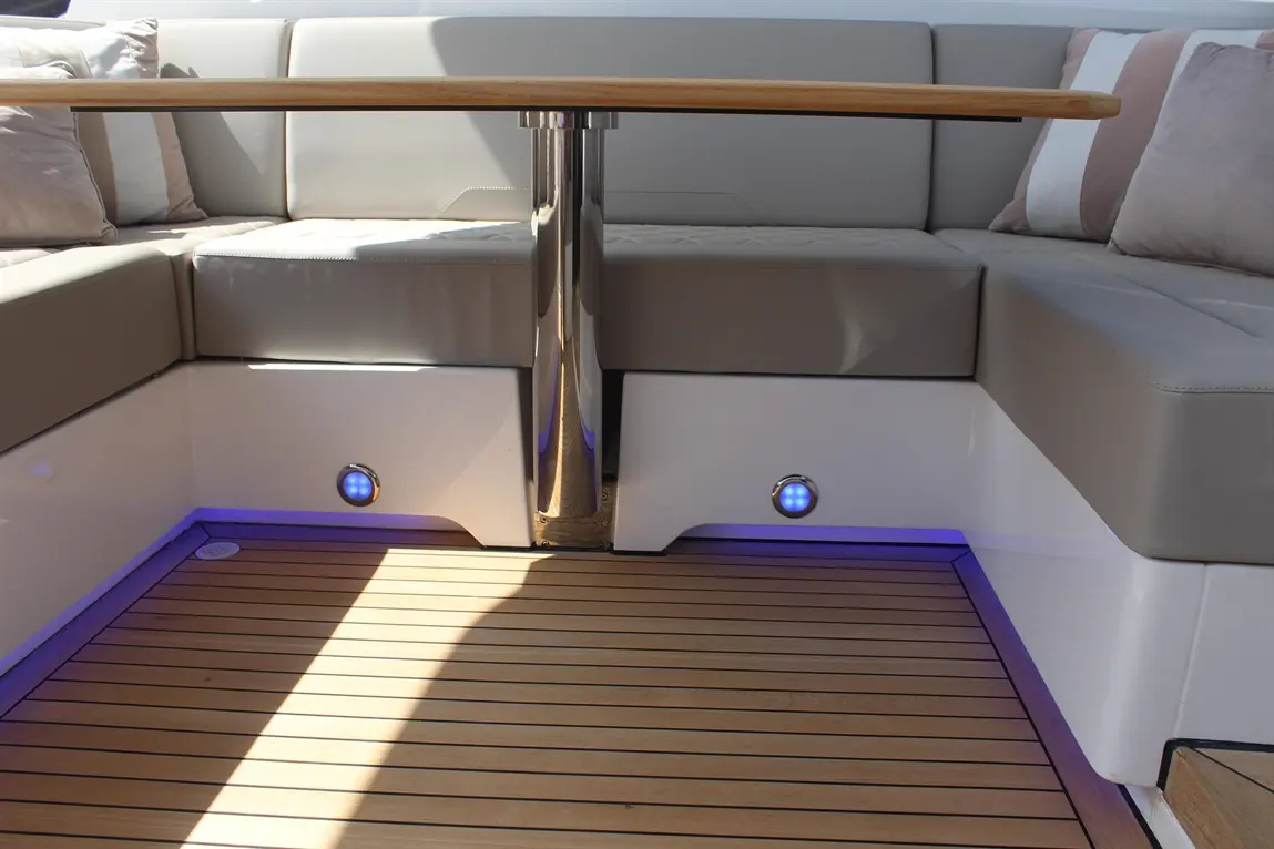Modern boat interior with a grey upholstered seating area, wooden table, and illuminated features in a sunny setting.