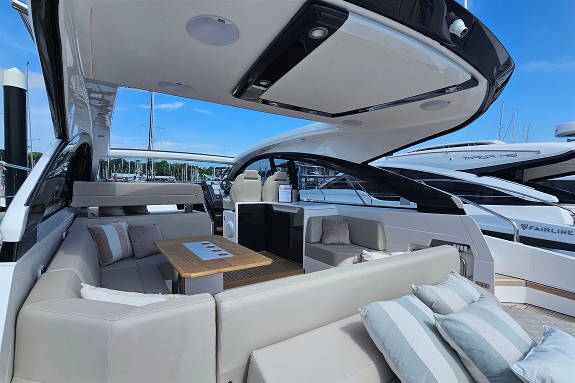 A modern yacht interior with seating, a wooden table, and a view of a marina under a clear blue sky. Visible text includes "FAIRLINE.