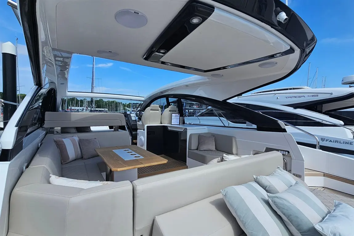 A modern yacht interior with seating, a wooden table, and a view of a marina under a clear blue sky. Visible text includes "FAIRLINE.