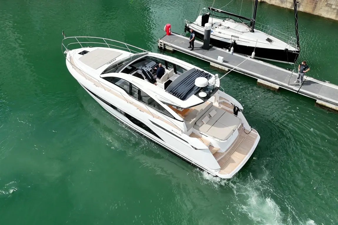 Luxury motor yacht manoeuvring in green waters near a marina, with crew members on the dock and a black sailboat in view.