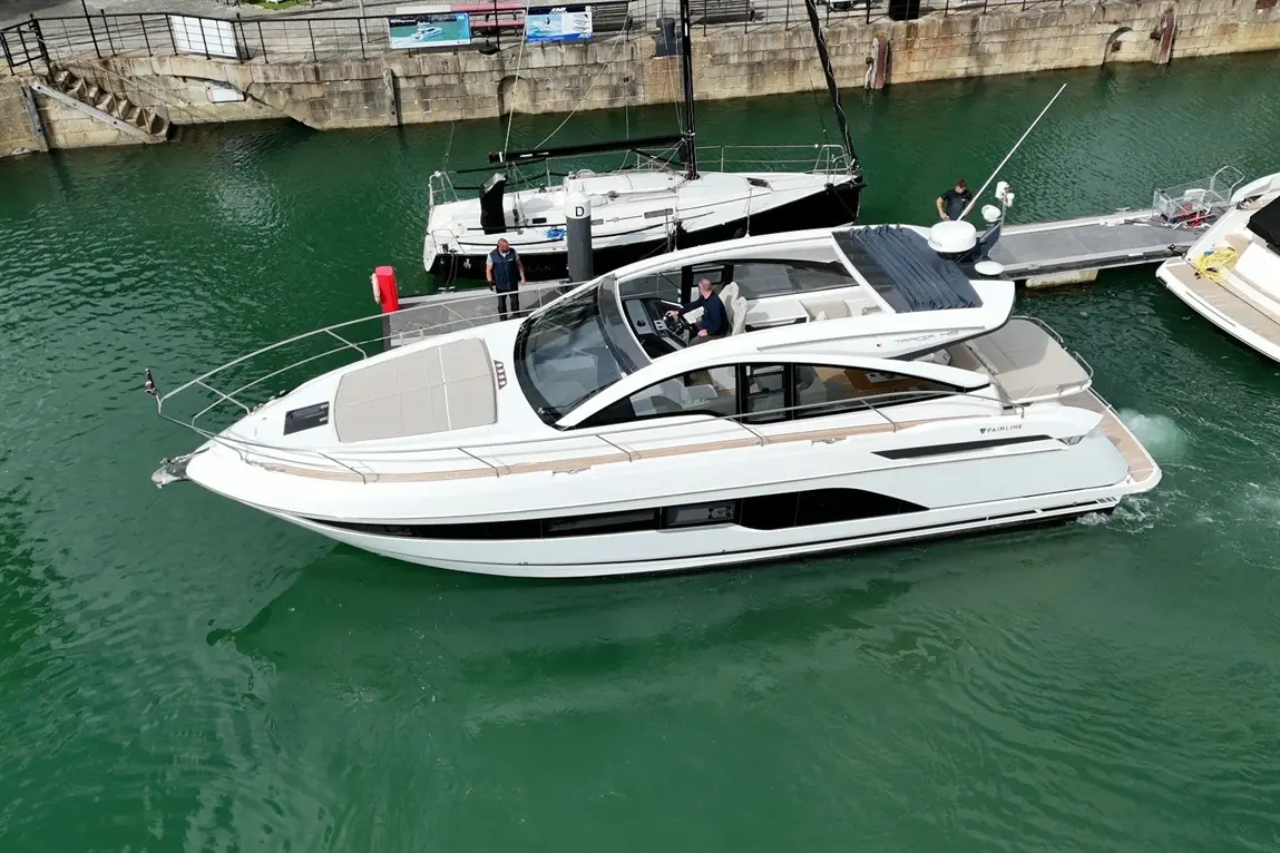 A sleek white motorboat navigates a green marina, with two crew members on board and a nearby docked yacht.