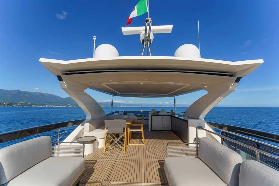 Luxury yacht interior with seating, dining area, and Italian flag, overlooking the ocean and mountains under a clear sky.