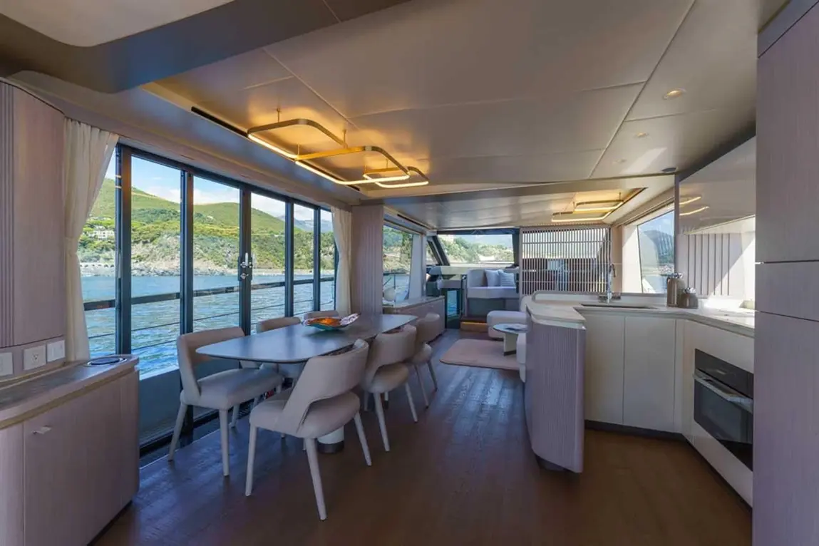 Bright, modern interior of a boat with dining area, large windows, and a sleek kitchen overlooking water and hills.