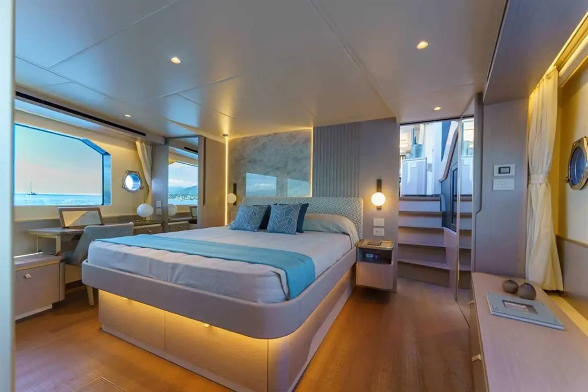 Modern yacht interior featuring a queen bed, desk, large windows, and stairs to upper levels, illuminated by soft lighting.