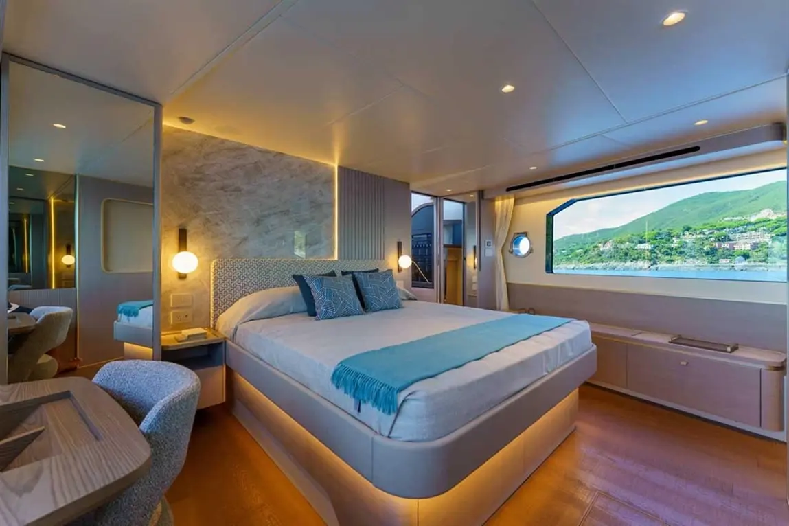 Modern bedroom with a large bed, decorative pillows, and a panoramic window overlooking green hills and water.