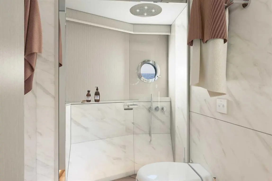Modern bathroom with marble walls, a circular window, shower, and sink, featuring brown towels and decorative toiletries.