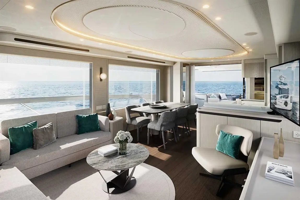 Stylish yacht interior featuring a lounge with seating, dining area, and kitchen, offering sea views through large windows.