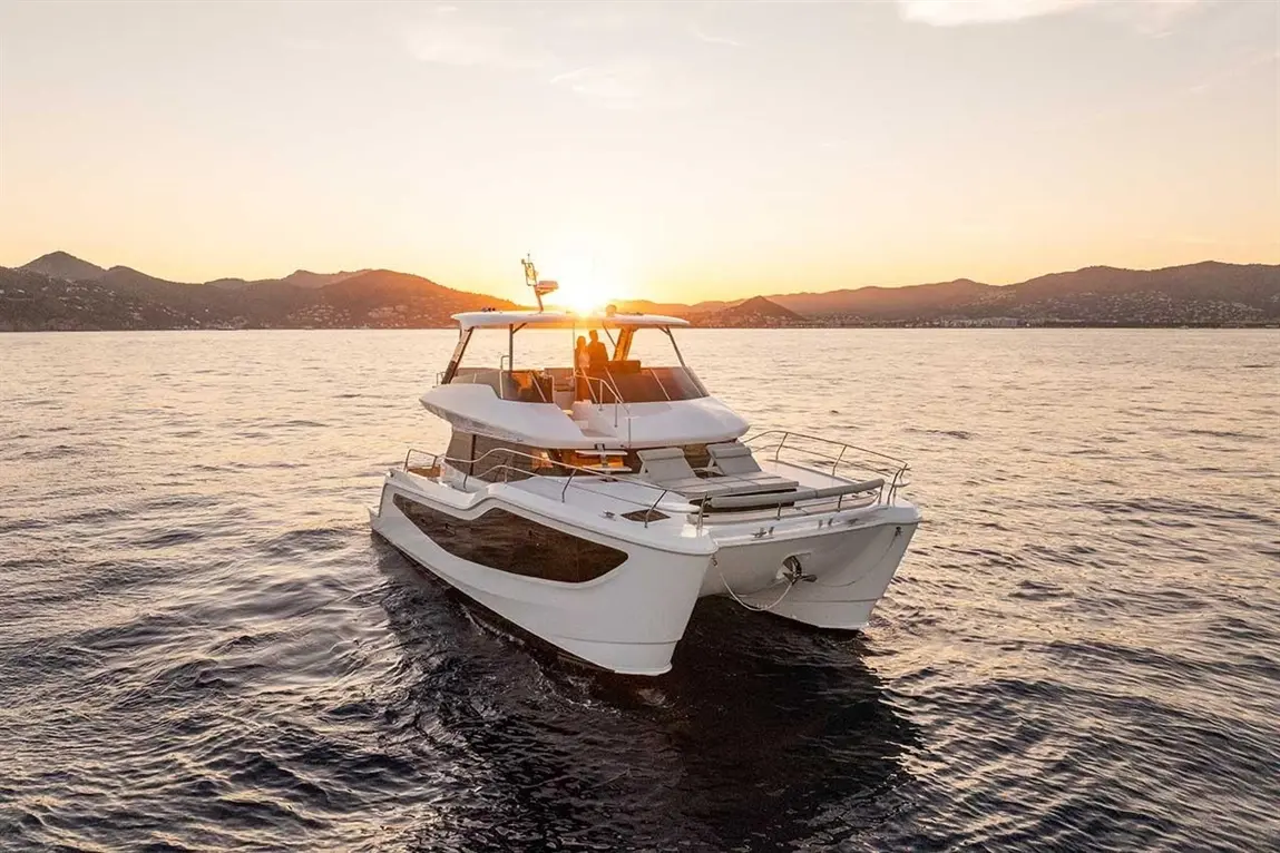 A sleek motor yacht sails on calm water at sunset, with mountains in the background and a warm glow illuminating the scene.