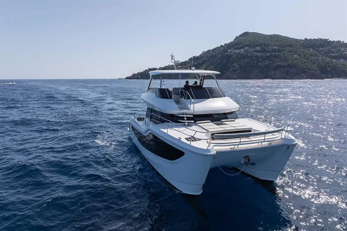 A sleek white catamaran cruising on calm blue waters, with a lush green hillside in the background.