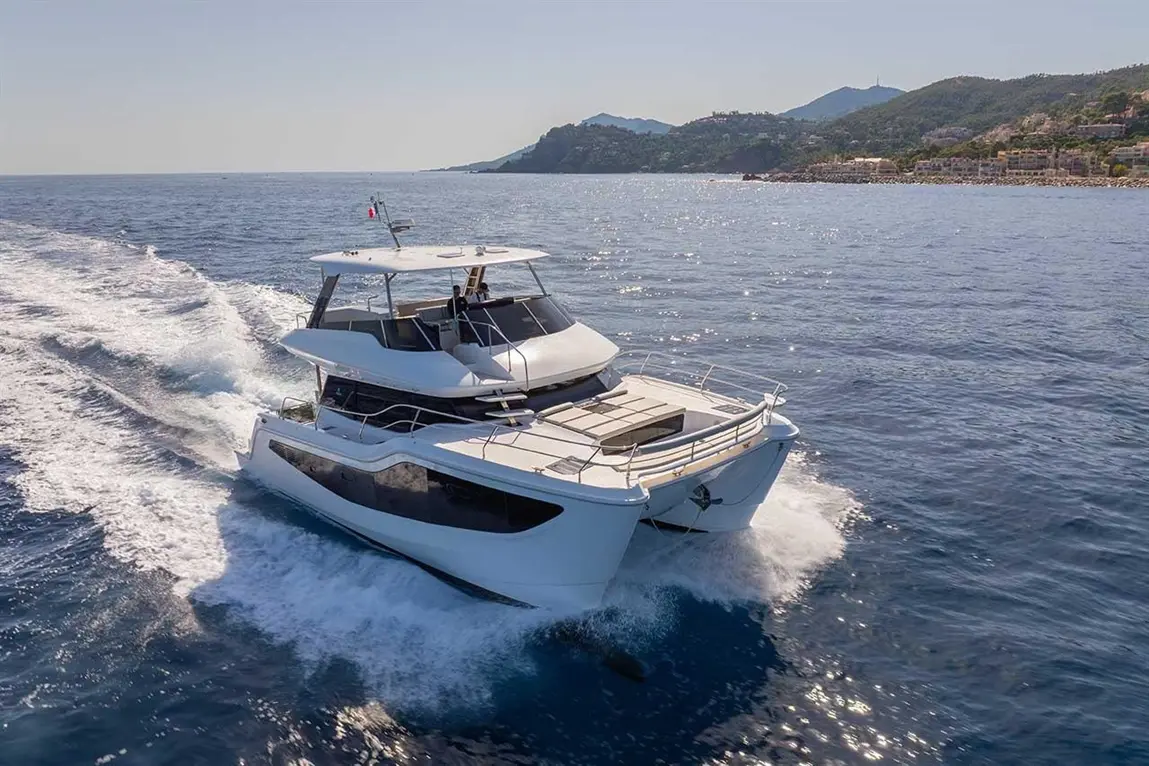 A luxury catamaran cruising on calm blue waters, leaving a white wake, with rocky coastline in the background.
