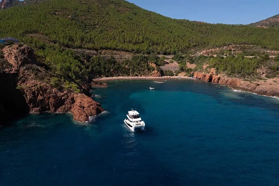 A luxury yacht sails near a secluded beach surrounded by lush greenery and rocky cliffs under a clear blue sky.