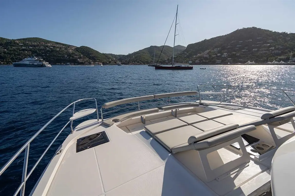 View from a yacht's deck with sun loungers, showcasing the marina, hills, and a sailing boat in the distance on calm waters.