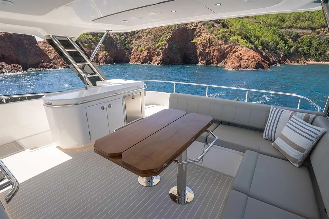 Luxury yacht deck featuring a wooden table, grey seating, and scenic views of rocky coastline and blue waters.