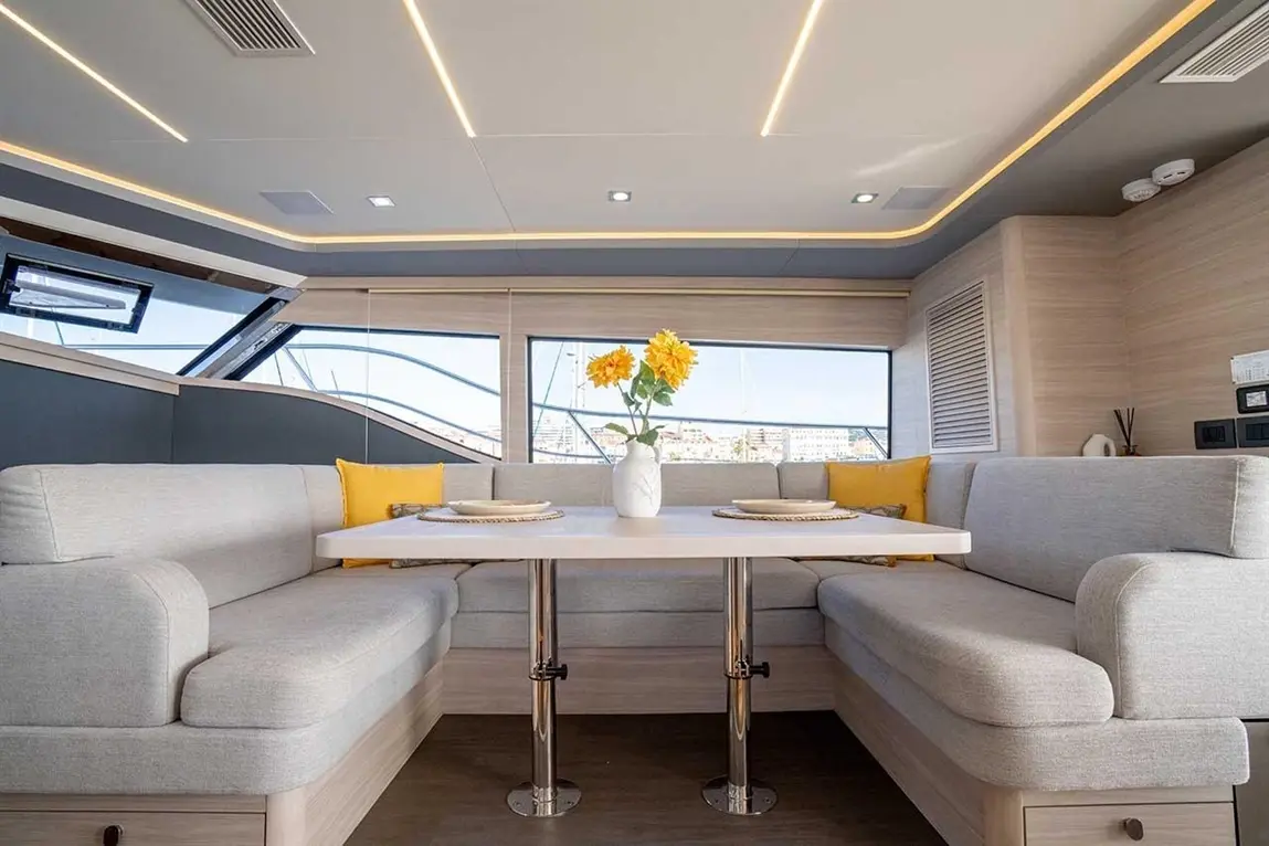 Modern yacht interior with a dining area, neutral sofas, a white table, and a vase of yellow flowers, overlooking a marina.