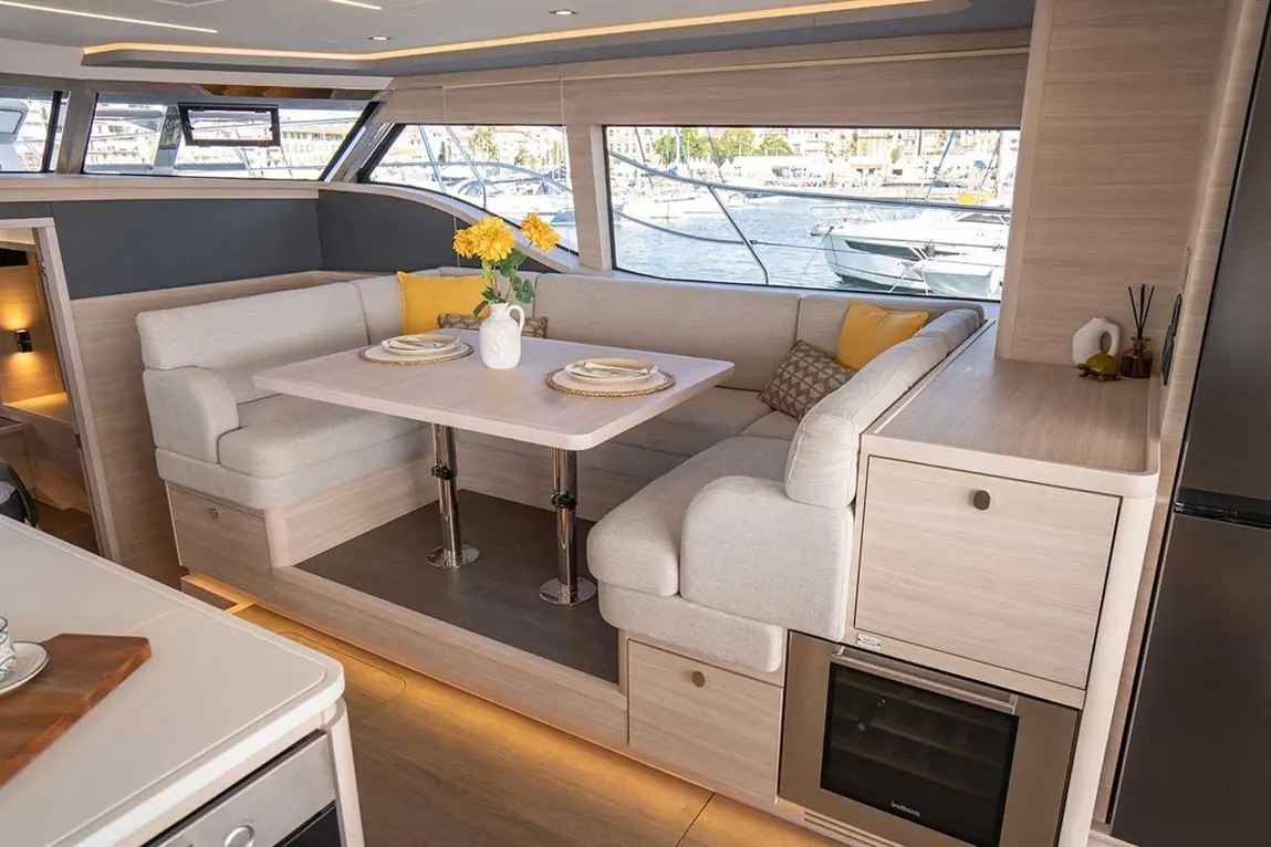 Modern boat interior featuring a dining area with a light wooden table, grey seating, flowers, and views of a marina.