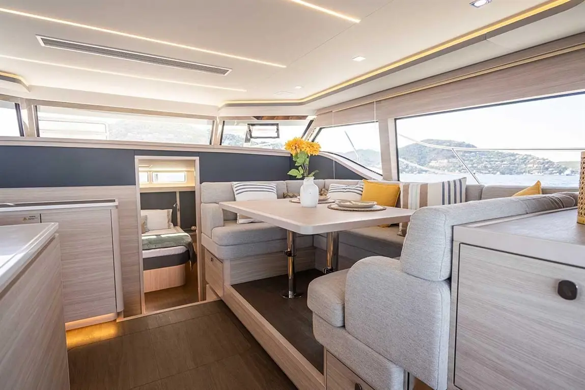 Modern yacht interior featuring a dining area, plush seating, a small kitchenette, and views of the sea through large windows.