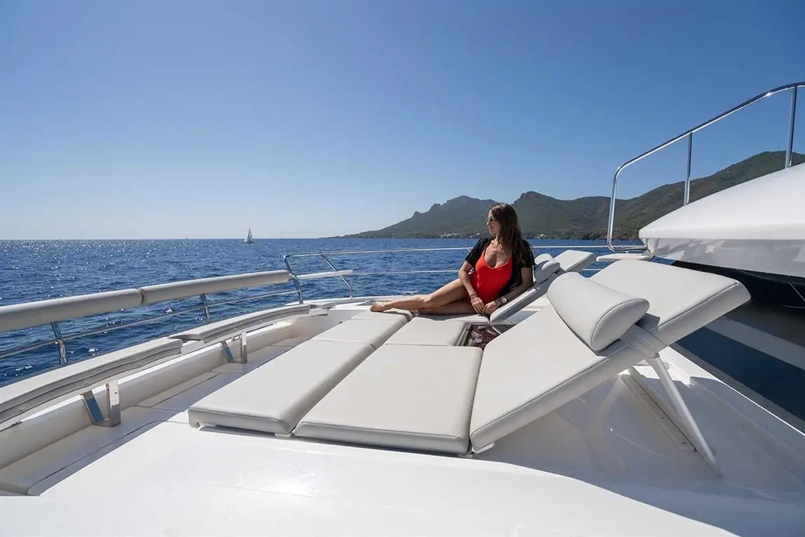 A woman in a red swimsuit relaxes on sun loungers aboard a yacht, with mountains and ocean in the background.
