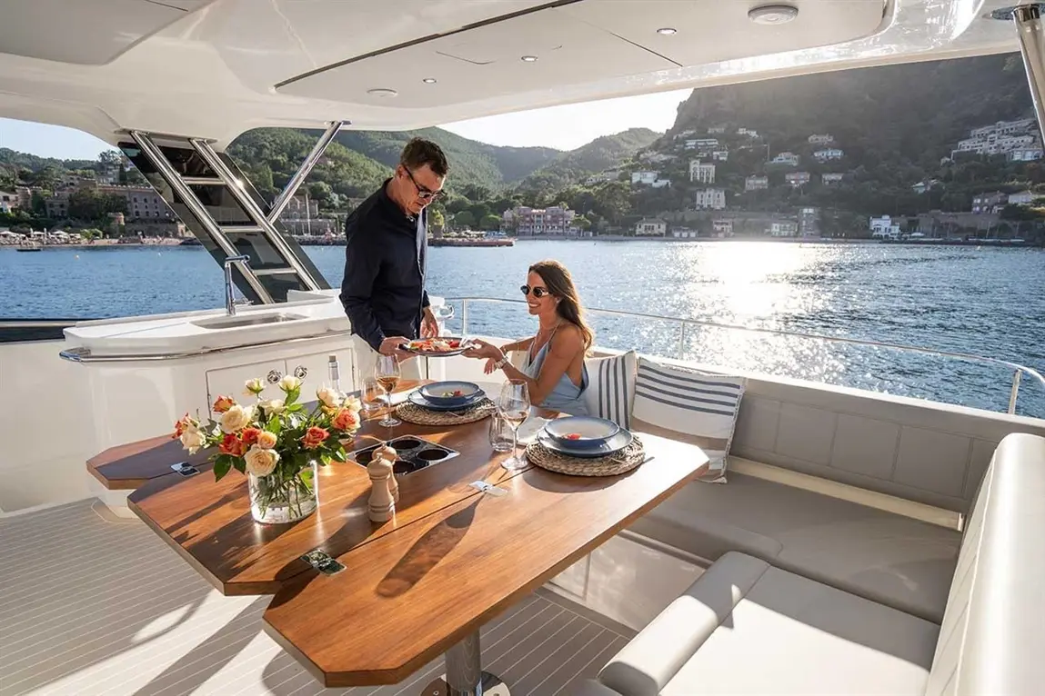 Couple dining on a yacht, enjoying a meal with flowers on the table and scenic coastal view in the background.
