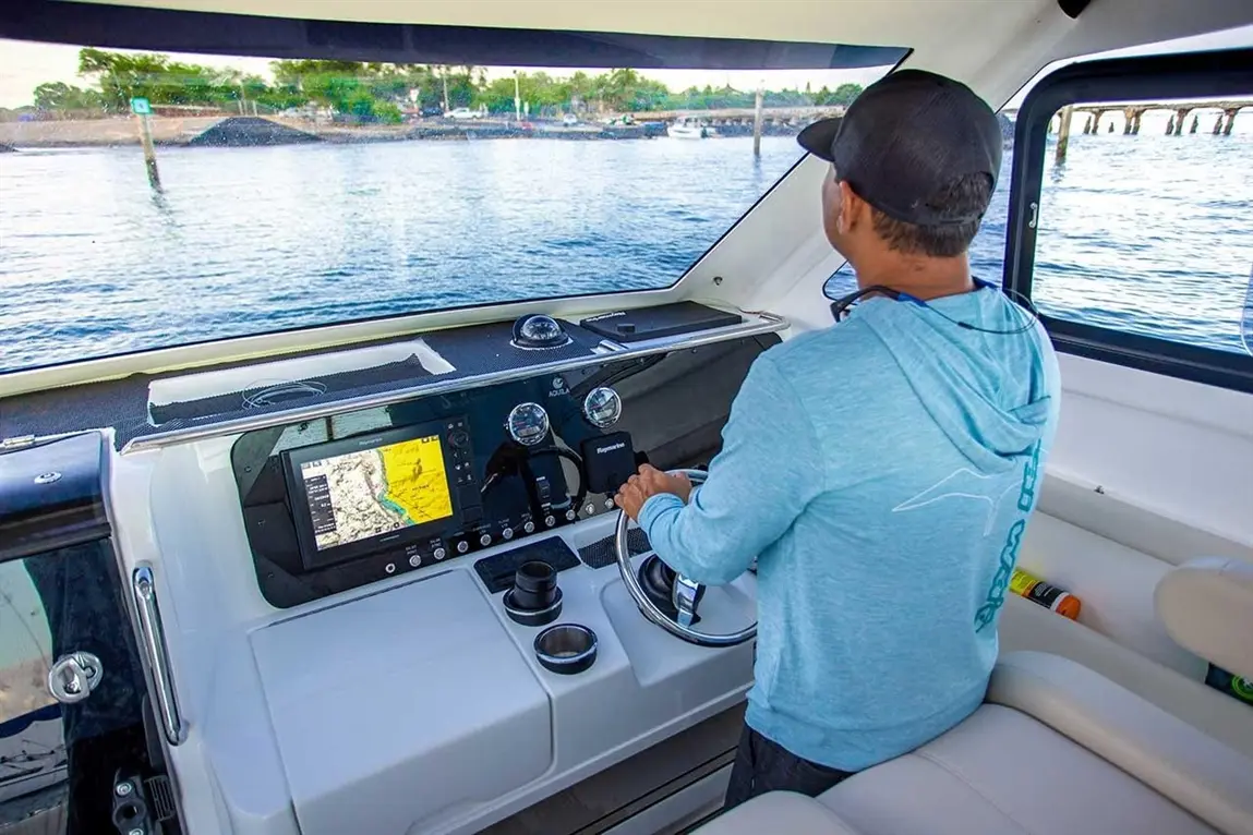 A man steering a boat at the control panel, featuring a chart display, with calm waters and greenery visible outside.