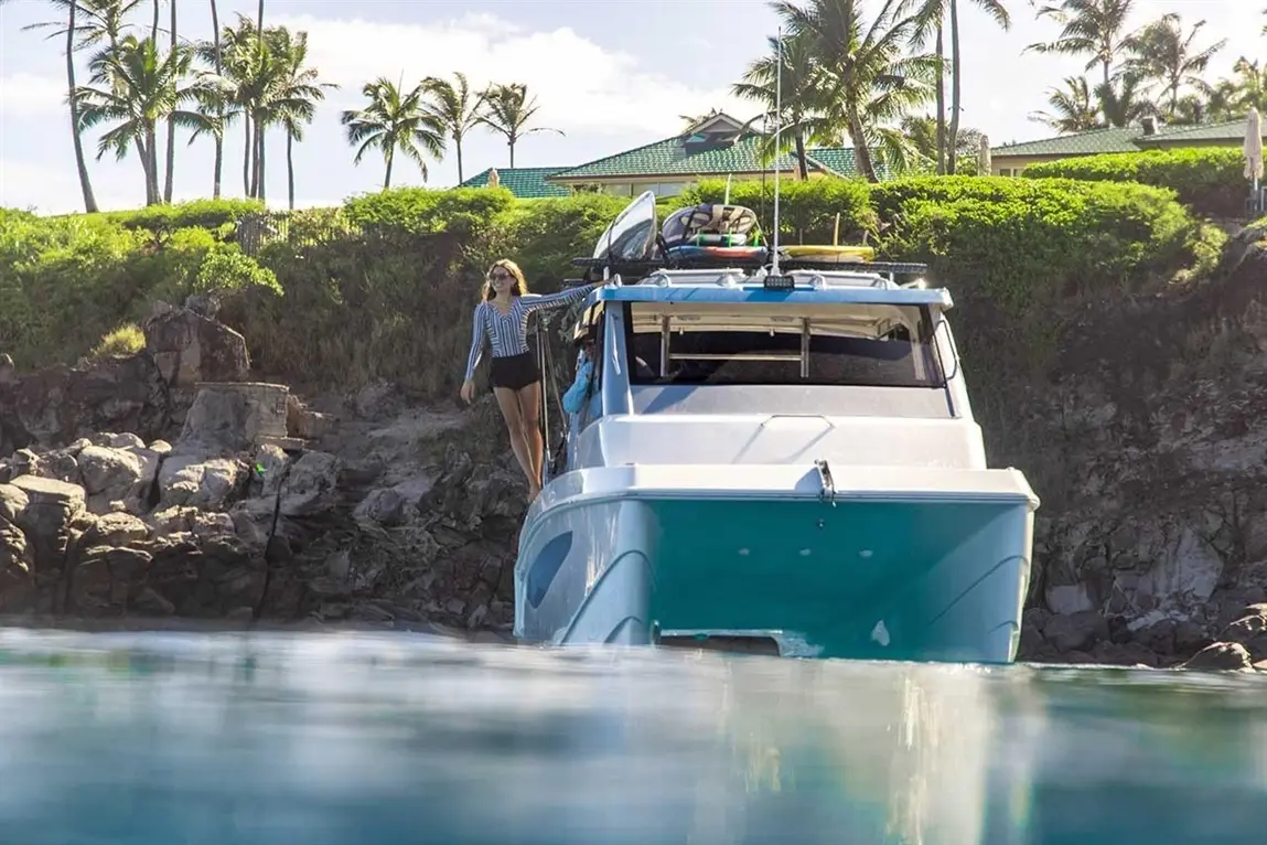 A woman in a striped long-sleeve shirt stands on a boat near rocky shore, palm trees and a green-roofed building in the background.