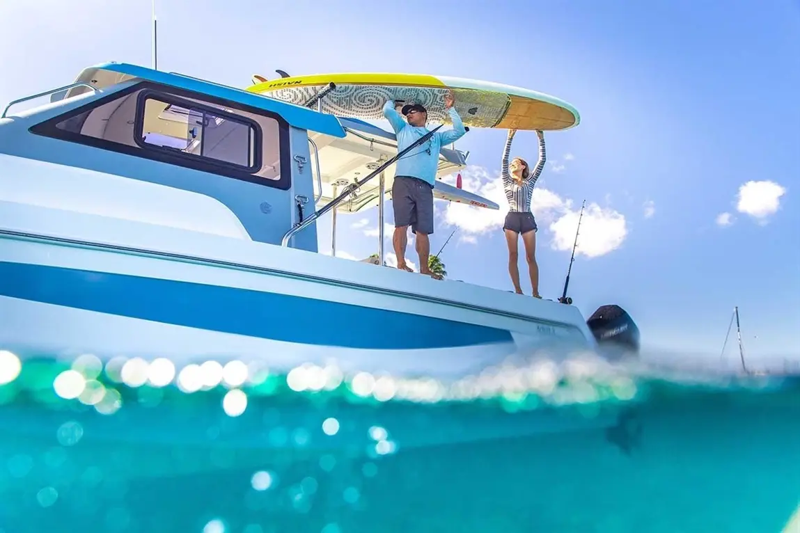 A man and woman on a boat prepare for paddleboarding, with a bright blue sky and water glistening below.