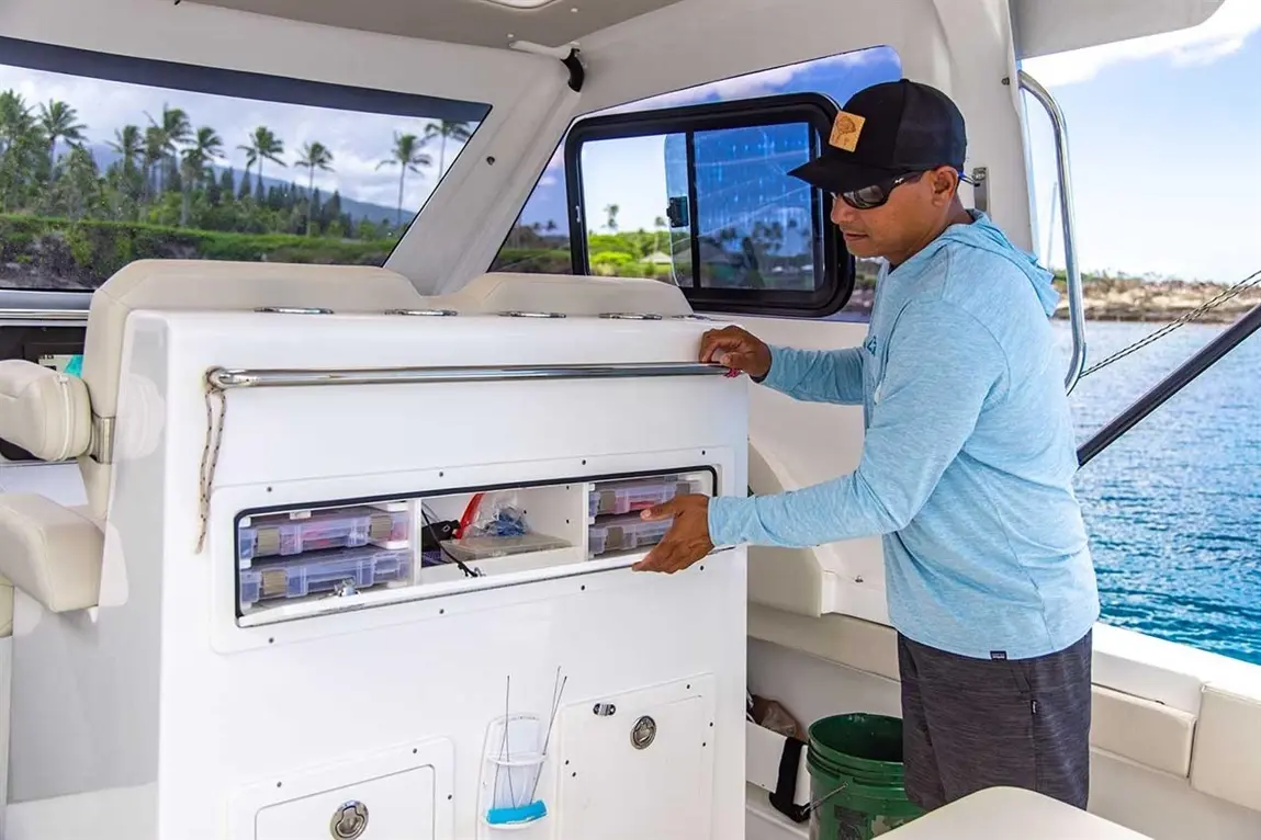Man in a blue hoodie accessing storage compartments on a boat, with tropical scenery visible through large windows.