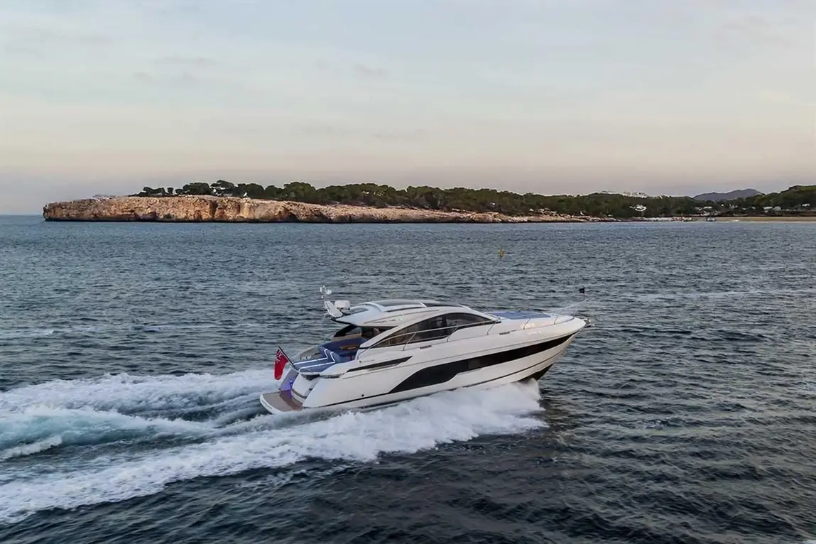A sleek motorboat speeds through calm waters near a rocky shoreline and greenery under a clear sky.