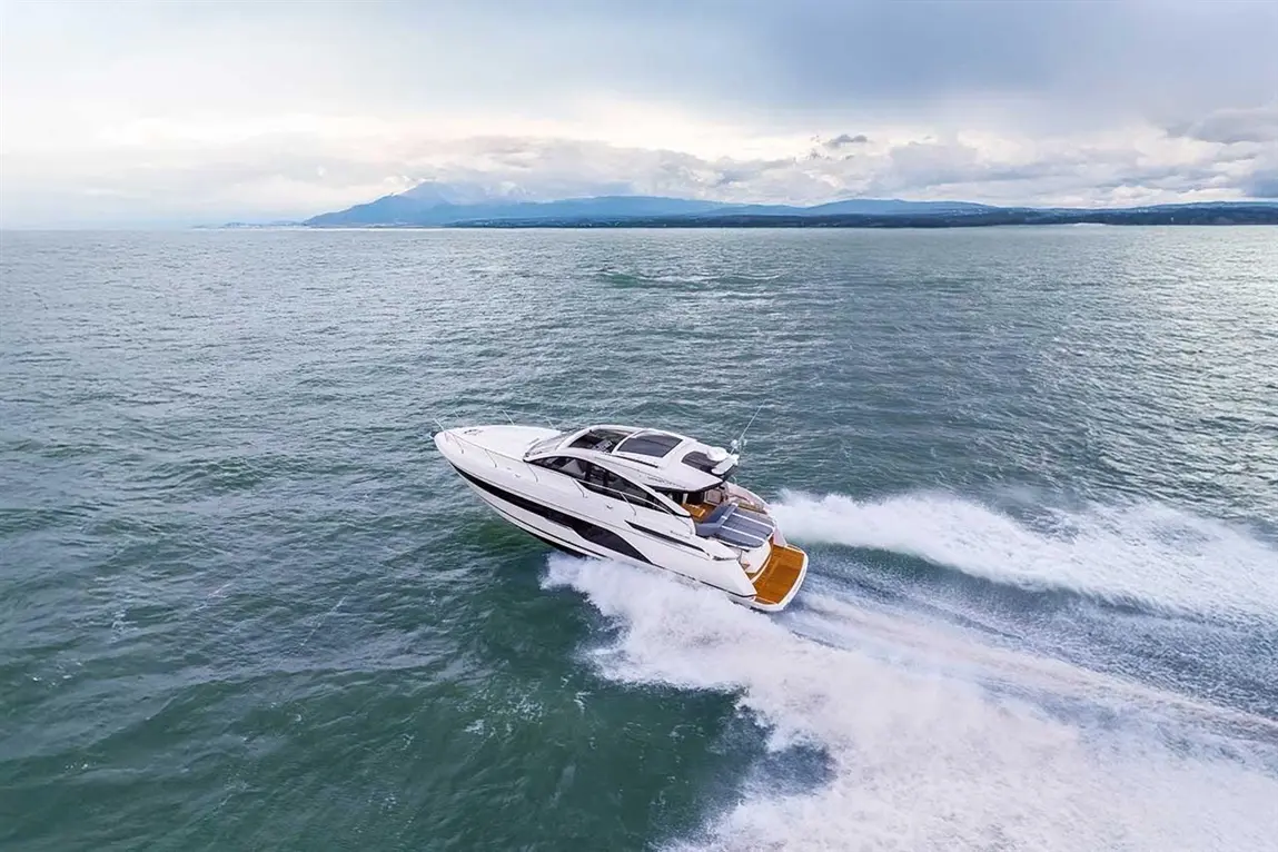 A luxury motorboat glides across the water, leaving a wake, with mountains and cloudy skies in the background.