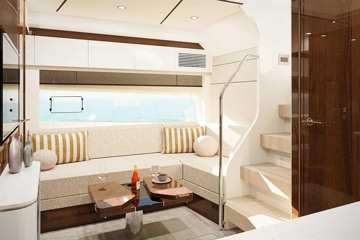 A stylish yacht interior featuring a sofa with striped cushions, a coffee table, and a staircase leading up, with sea views outside.