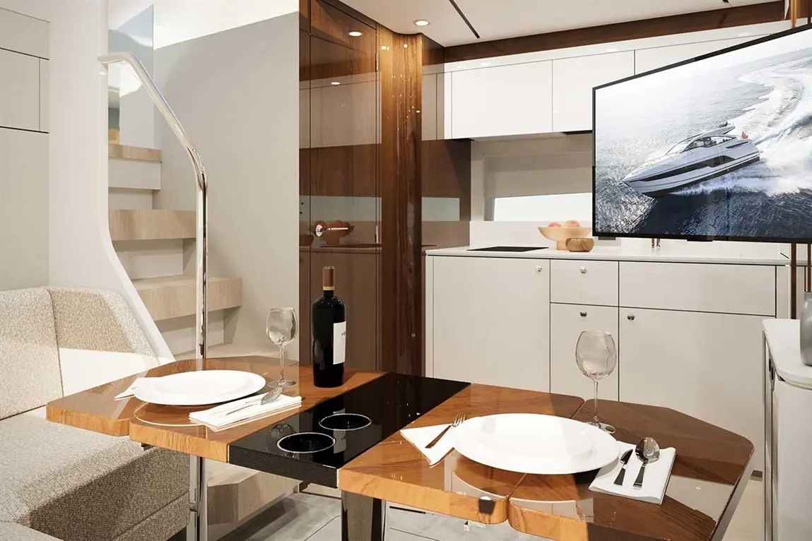 Modern dining area with a wooden table, wine, and a TV showing a luxury yacht, adjacent to a small kitchen.