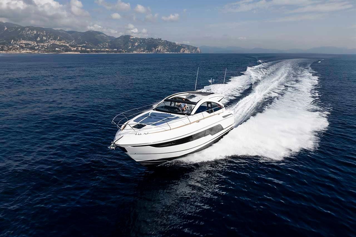 Luxury motorboat gliding through clear blue water, leaving a white wake; scenic mountains in the background under a cloudy sky.