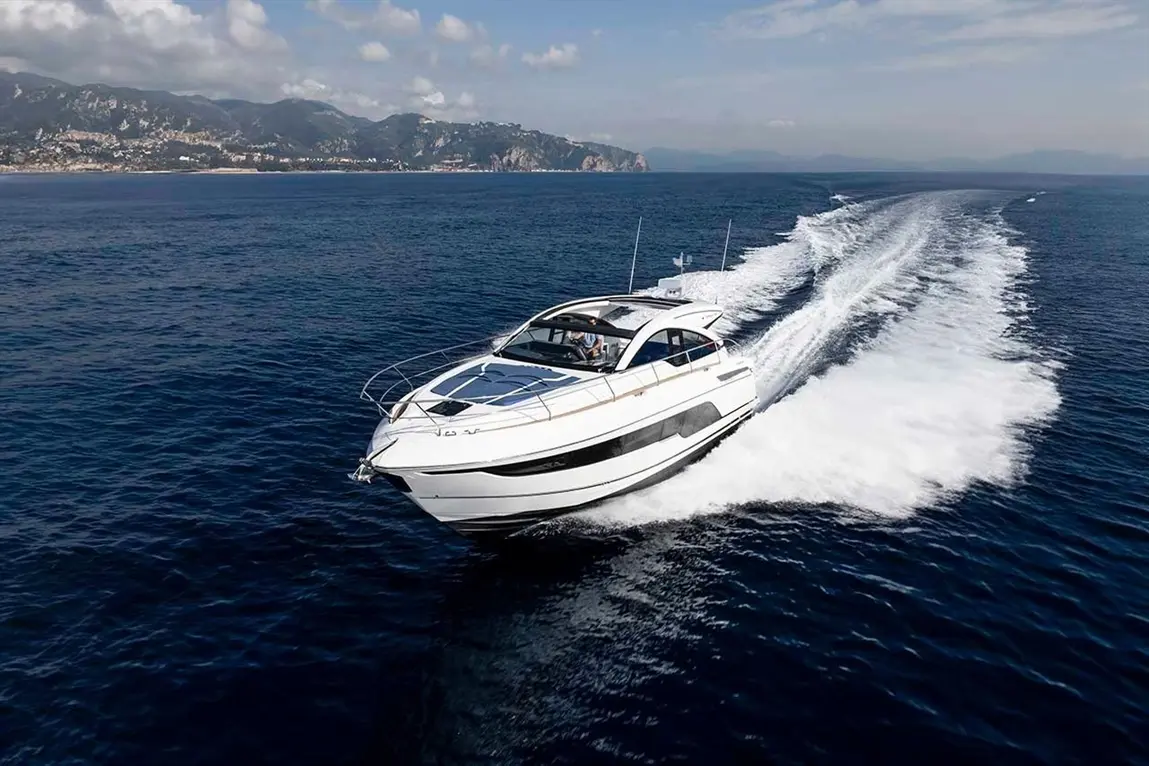 Luxury motorboat gliding through clear blue water, leaving a white wake; scenic mountains in the background under a cloudy sky.