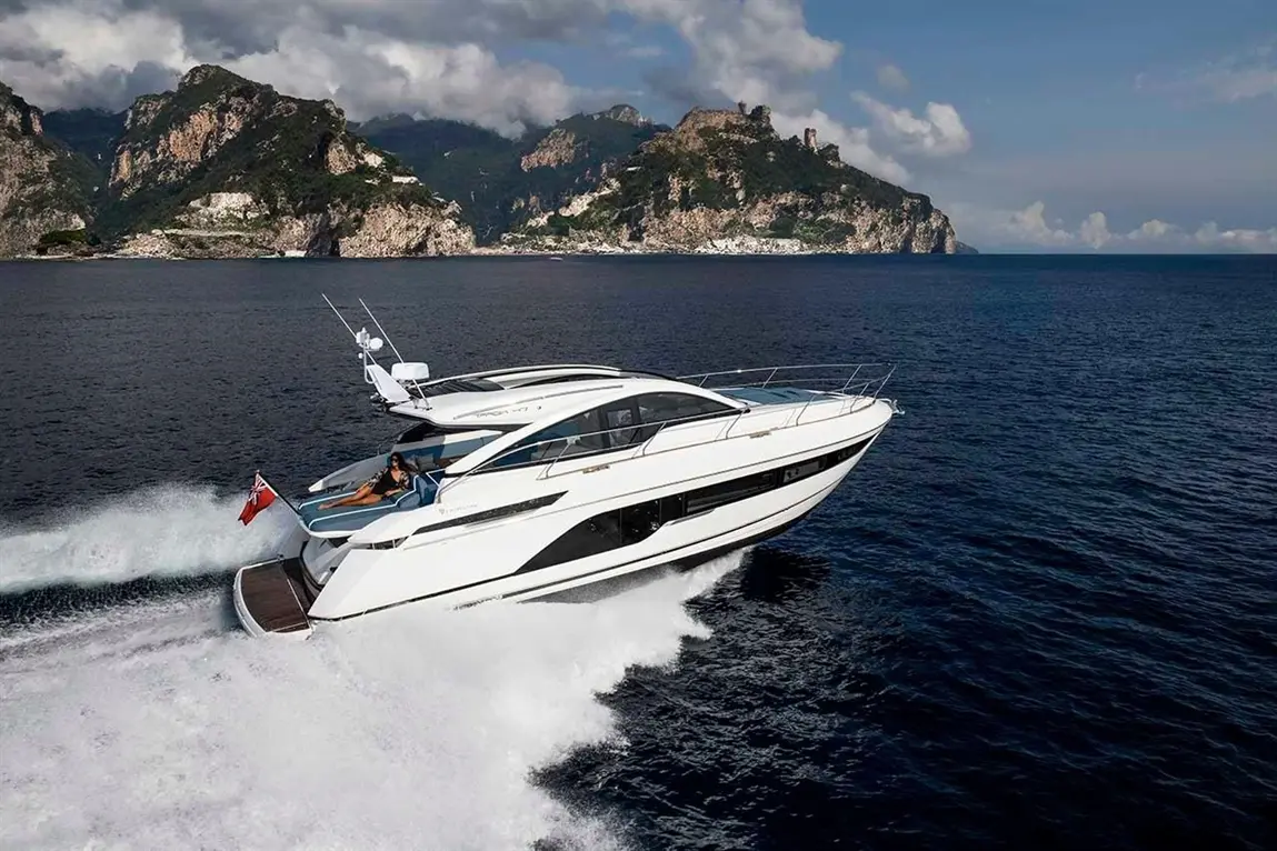 A sleek white luxury yacht speeds through azure waters, with dramatic rocky cliffs in the background under a cloudy sky.